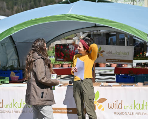 Zwei Frauen im Gespräch vor einem Messestand mit Pflanzen, Banner wild und kultiviert, Inklusion